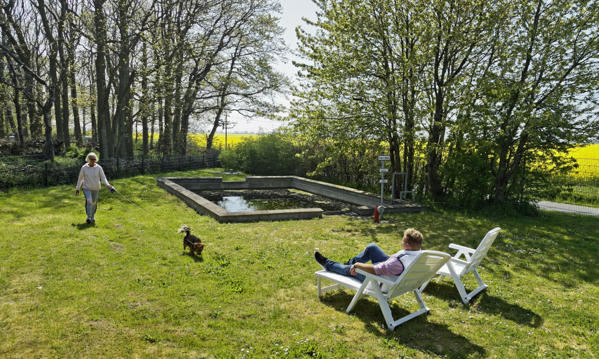 At KNAUS Campingpark Rügen, a woman strolls with her dog across a grassy meadow, while a man unwinds on a lounge chair beside a small pond encircled by trees.
