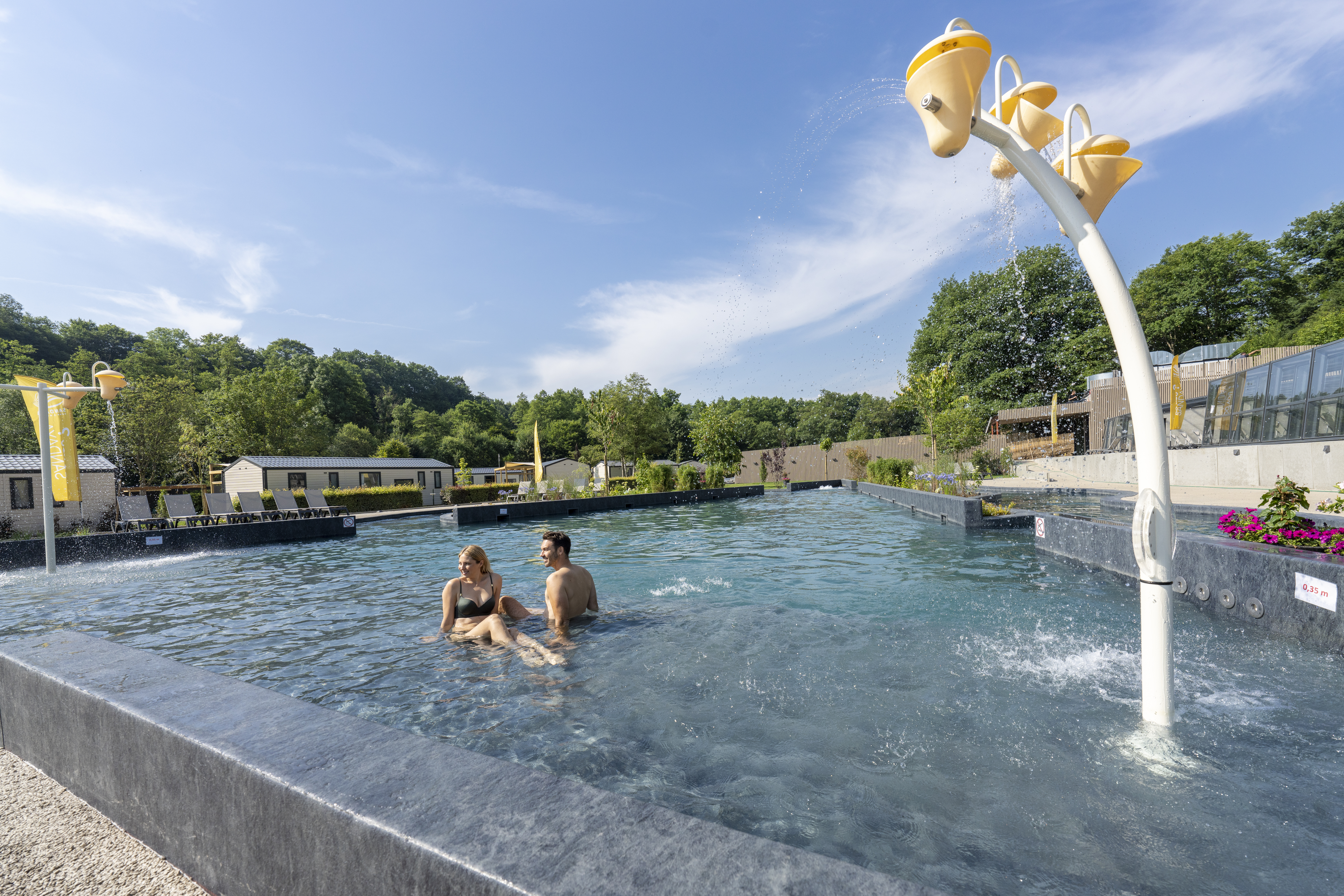 At Parc La Clusure, two individuals unwind in a shallow swimming pool adorned with playful water elements, encircled by vibrant foliage and cabins.