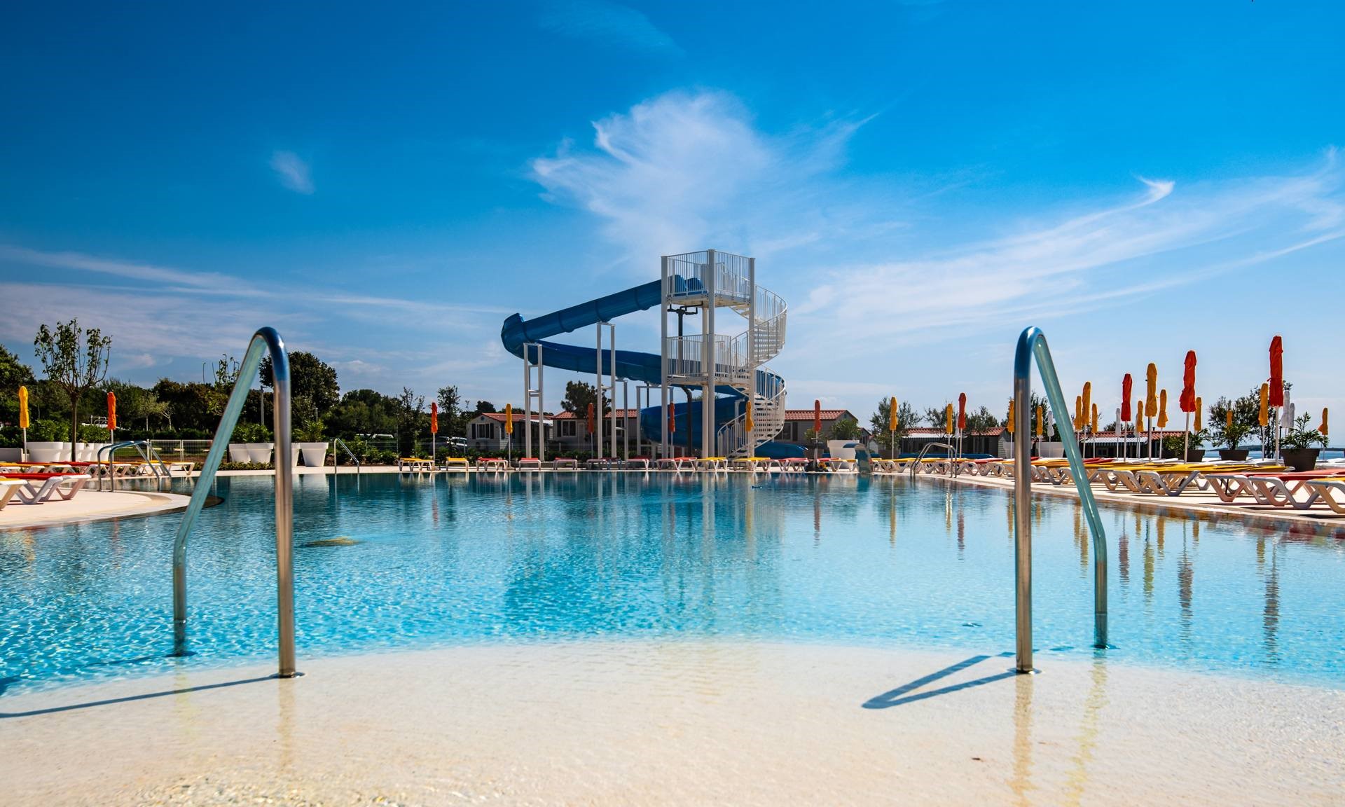 At Grand Kazela, a water slide stretches over a spacious swimming pool encircled by lounge chairs and umbrellas beneath a clear blue sky.