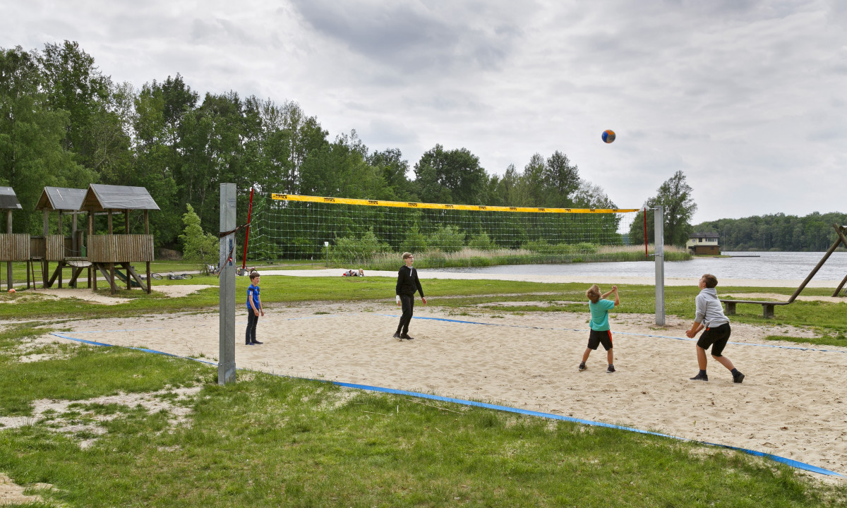 At KNAUS Campingpark Oyten, children are enjoying volleyball on a sandy court beside a lake, surrounded by lush trees and wooden structures nearby.