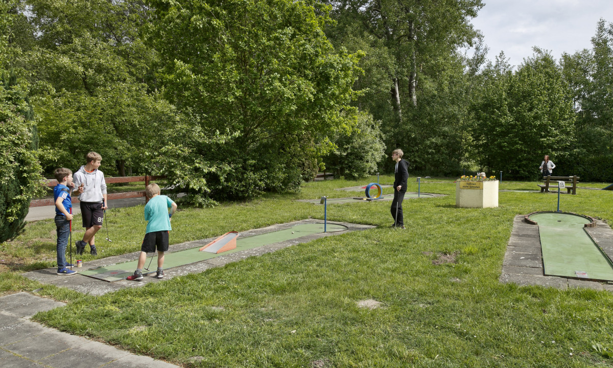 At KNAUS Campingpark Oyten, individuals are enjoying mini-golf on a grassy course encircled by trees in a park-like environment.