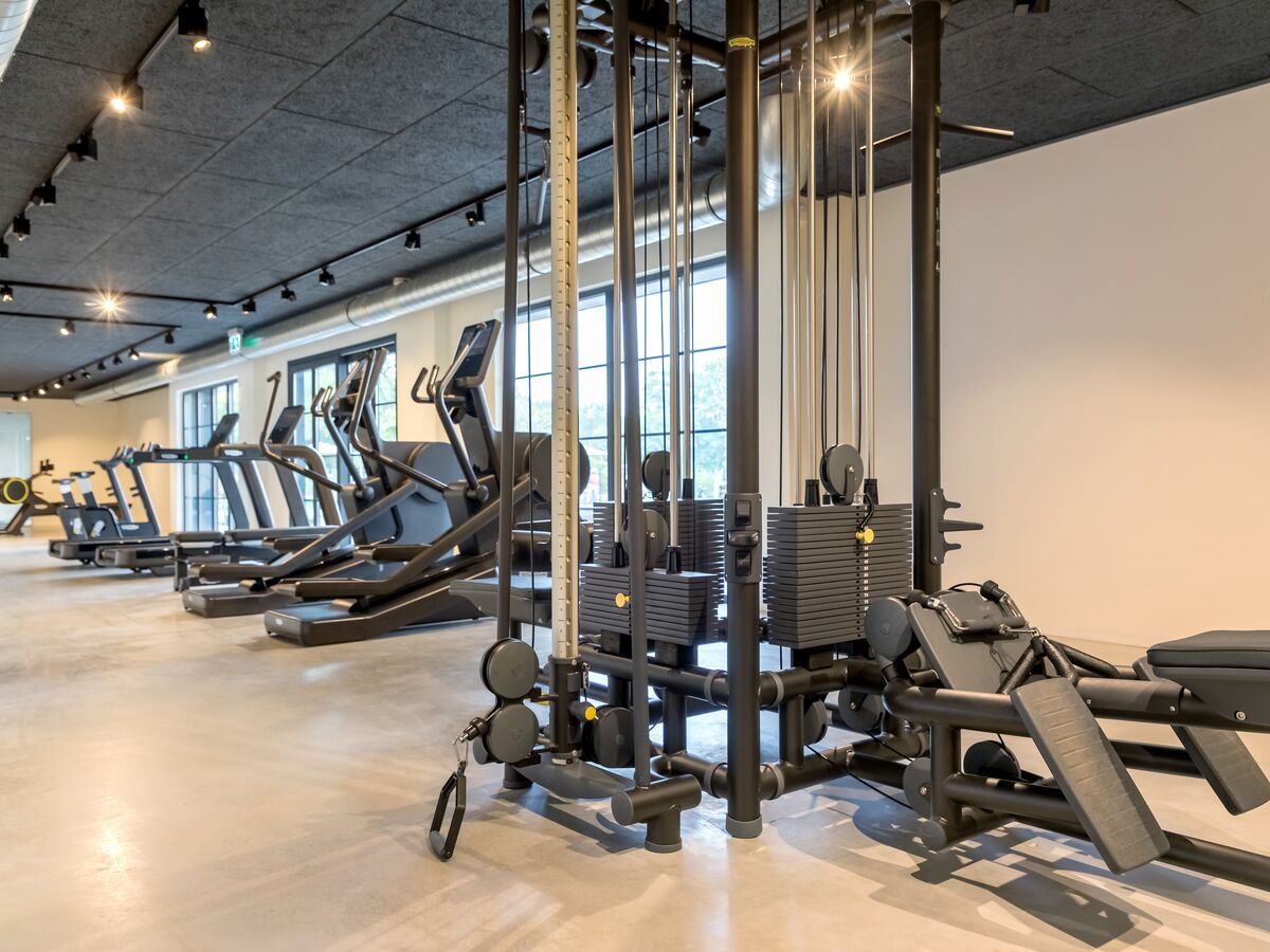 At De Twee Bruggen, exercise equipment is prepared in a tidy, brightly lit fitness center featuring expansive windows and a contemporary industrial style.