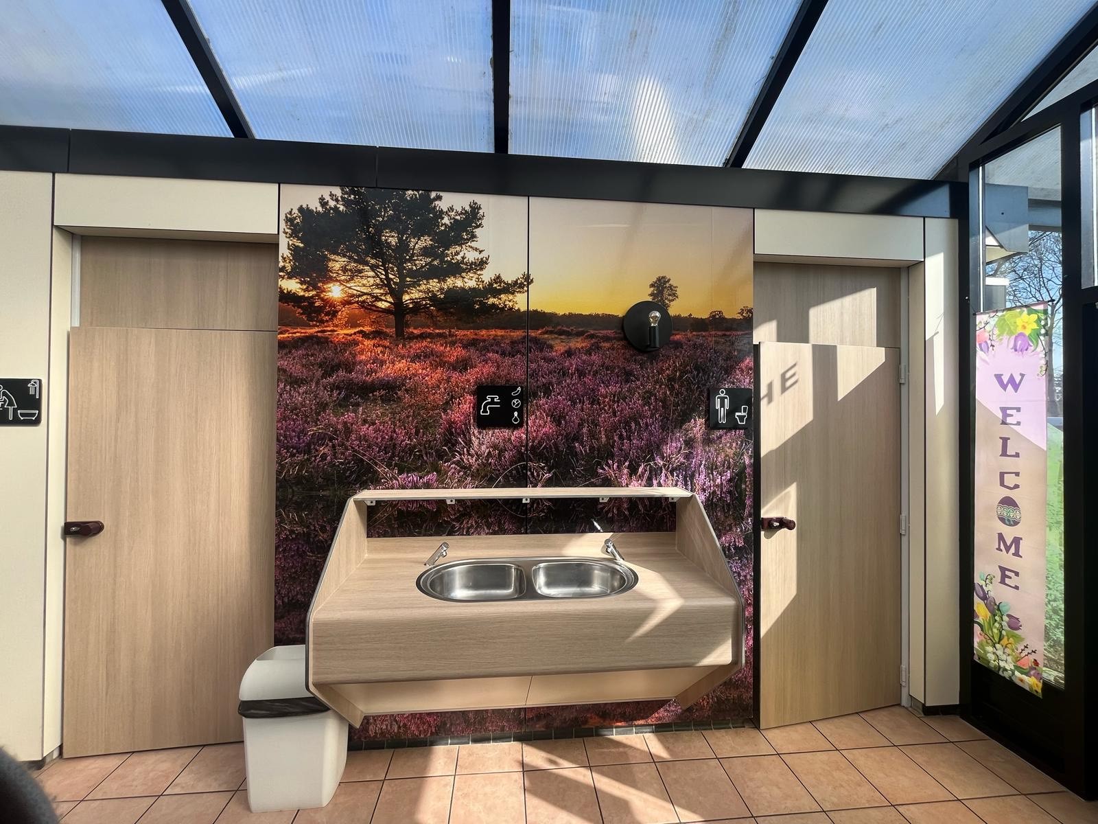 At de Vinkenkamp, there is a sink area adorned with a picturesque wallpaper backdrop, bordered by two restroom entrances within a brightly lit restroom space.