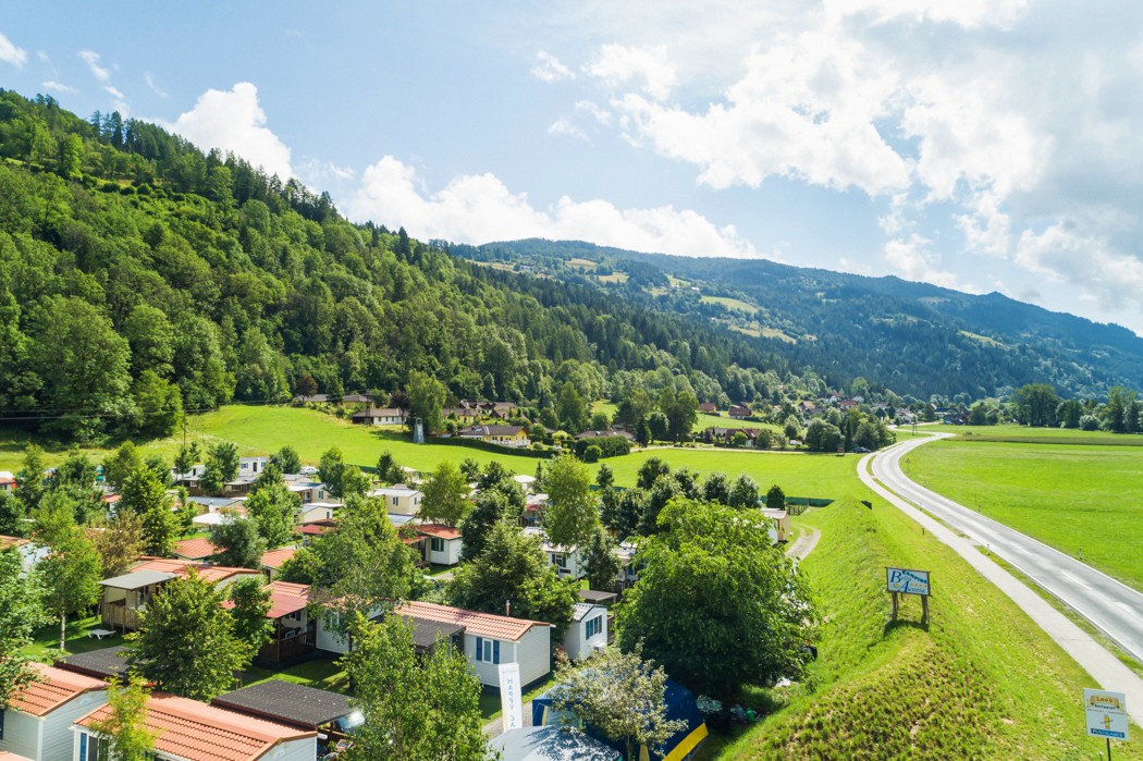 At Bella Austria, caravans are stationed amidst trees and grassy meadows, with wooded hills and a clear sky in the backdrop.