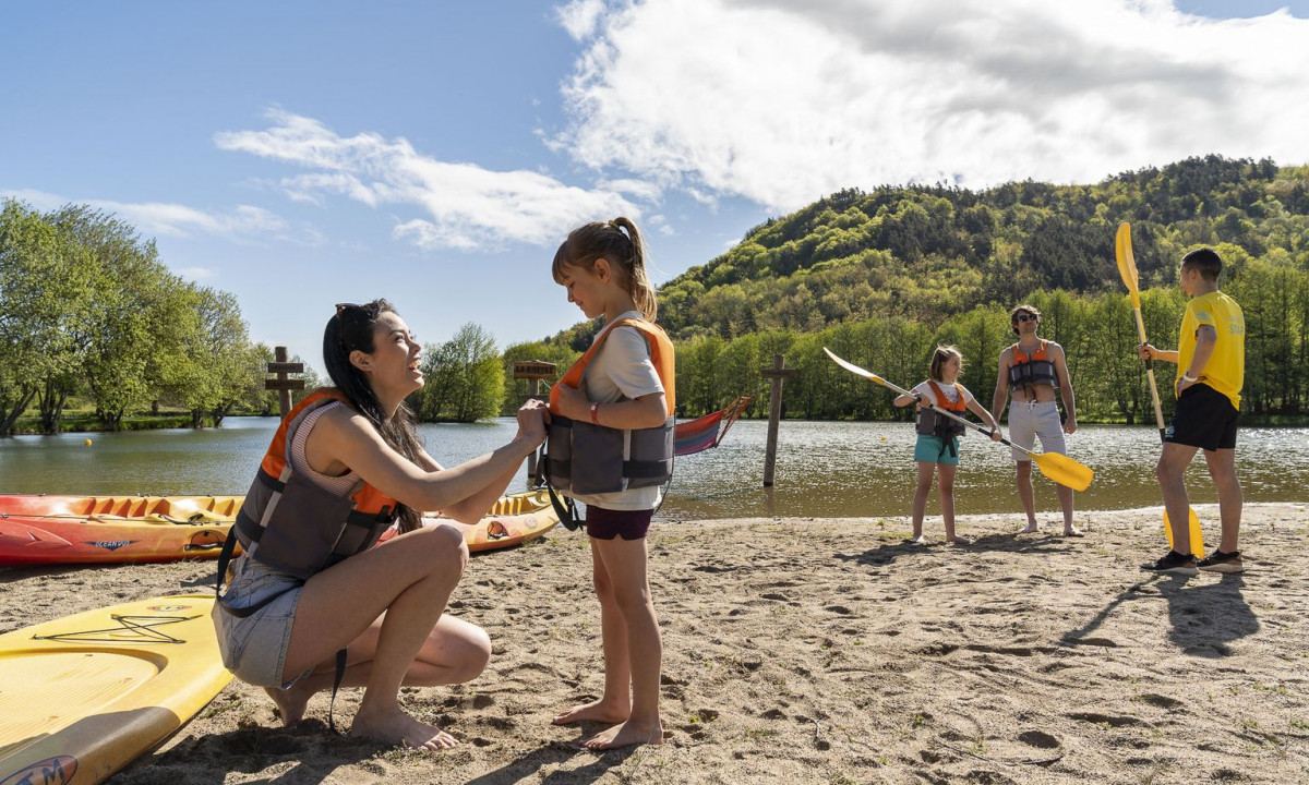 Woman adjusting child's life vest, others holding paddles, sandy beach by a lake, forested hills in the background at La Ribeyre