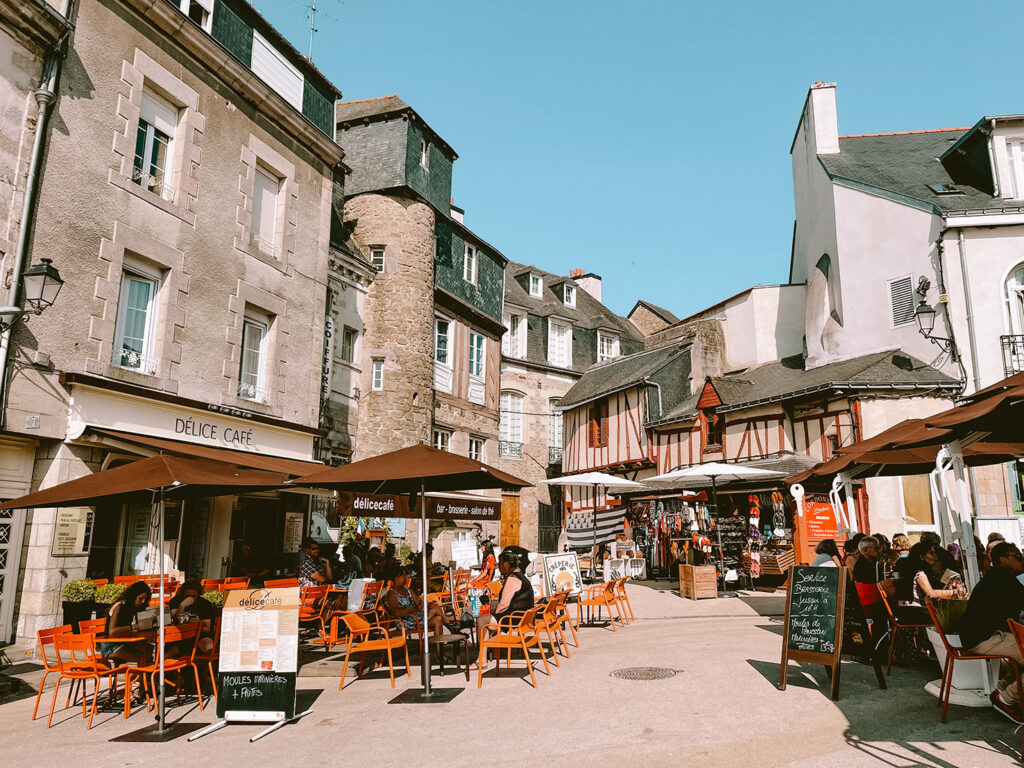 Café patrons eating and talking at outdoor tables amidst historic stone buildings and boutiques at Ker Goh Lenn