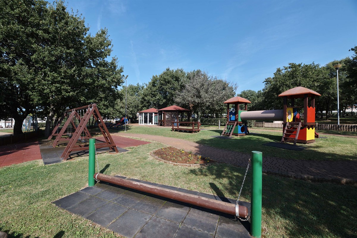 At Naturist Ulika, there is a playground with swings, vibrant play structures, and walkways encircled by grass and trees.