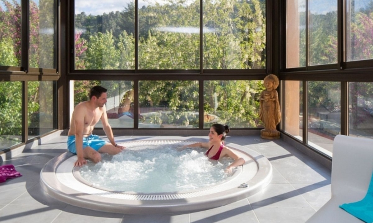At Les Ranchisses, two individuals are unwinding in a hot tub within a brightly lit room featuring expansive windows that provide a view of lush greenery.