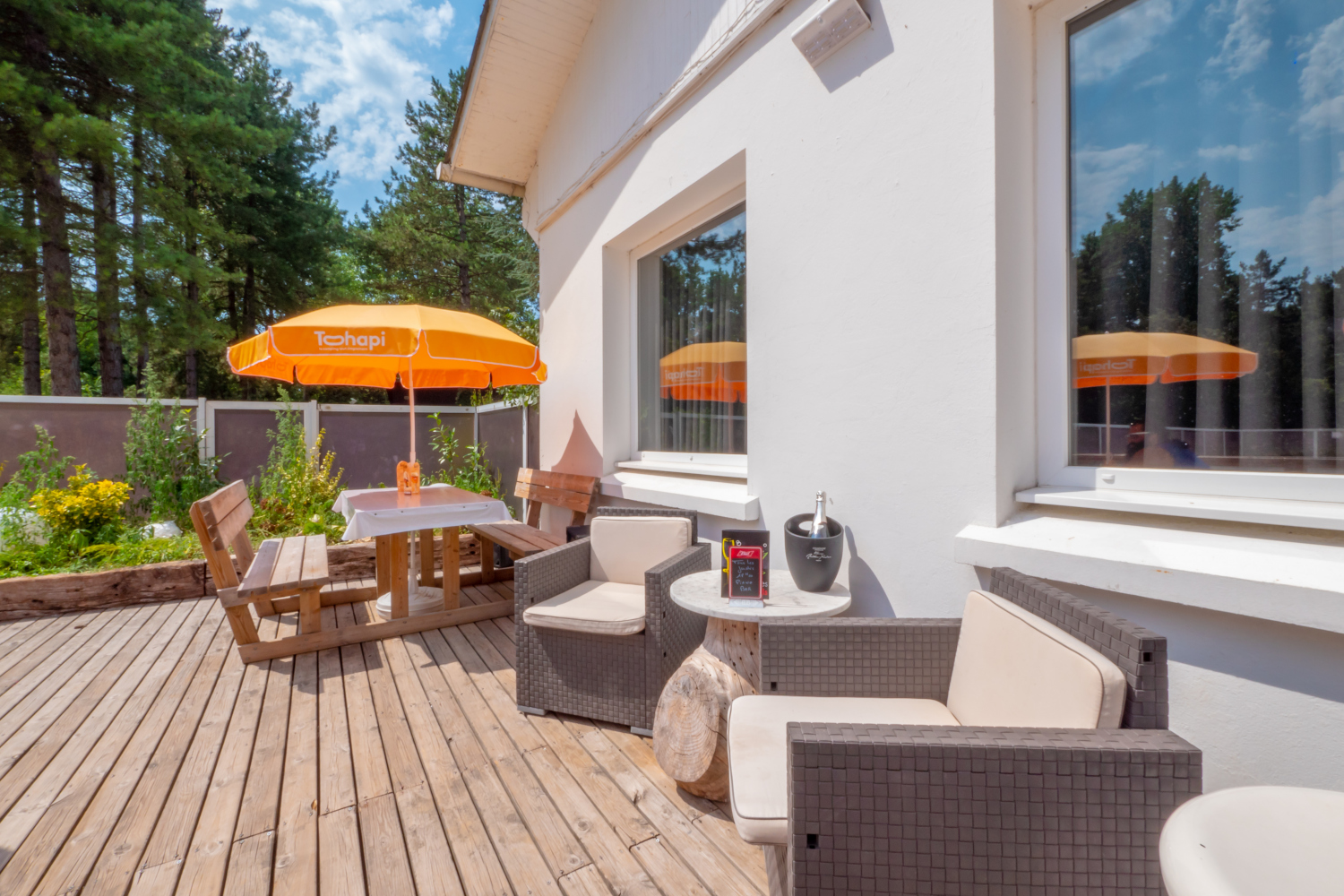 At La Dune Blanche, there is a patio featuring a wooden table, benches, and an orange umbrella next to a white house, encircled by lush greenery.