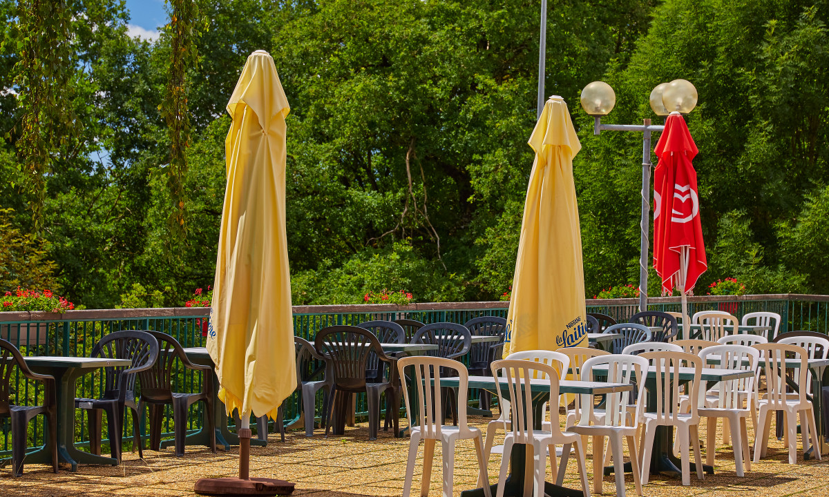 At Terrasses du Lac, collapsed umbrellas are positioned between lines of vacant plastic seats and tables on a terrace, encircled by vibrant foliage.