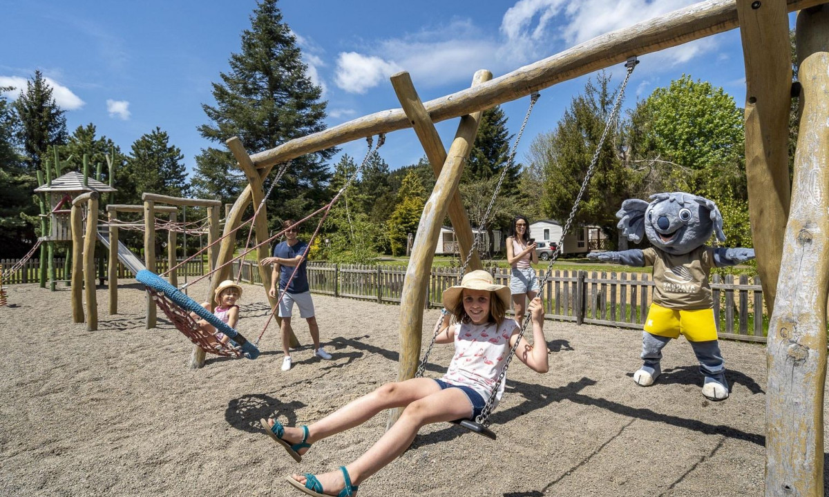 Children and a costumed character swinging in a playground surrounded by trees and cabins at La Ribeyre