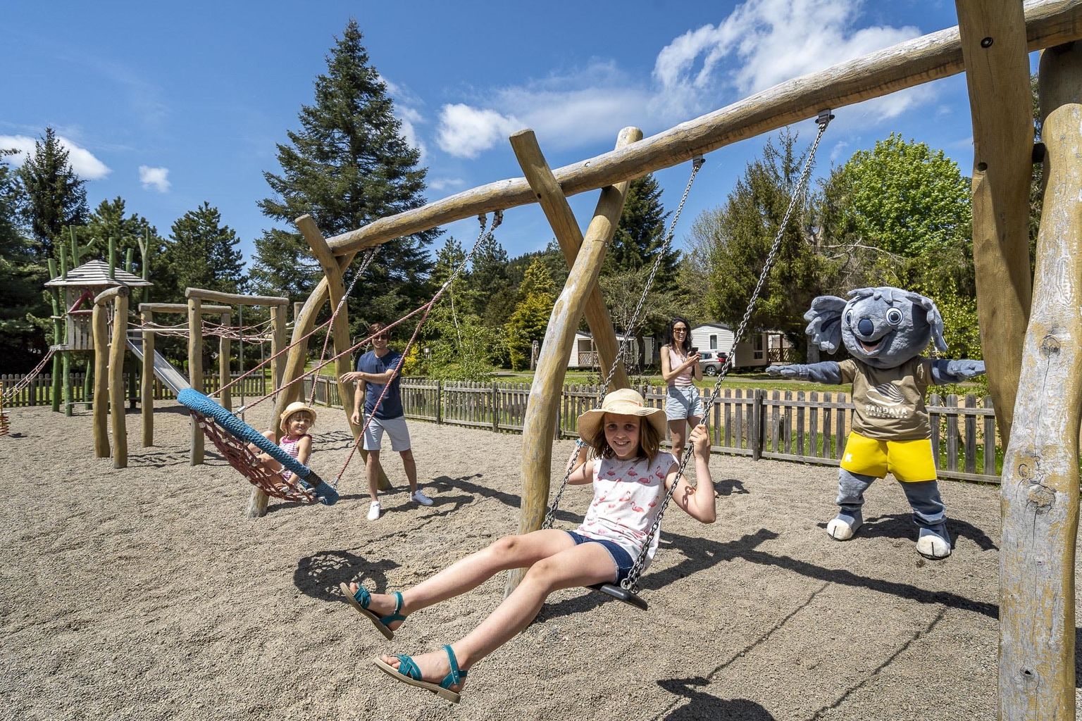 At La Ribeyre, kids and a costumed character are swinging in a play area encircled by trees and cabins.