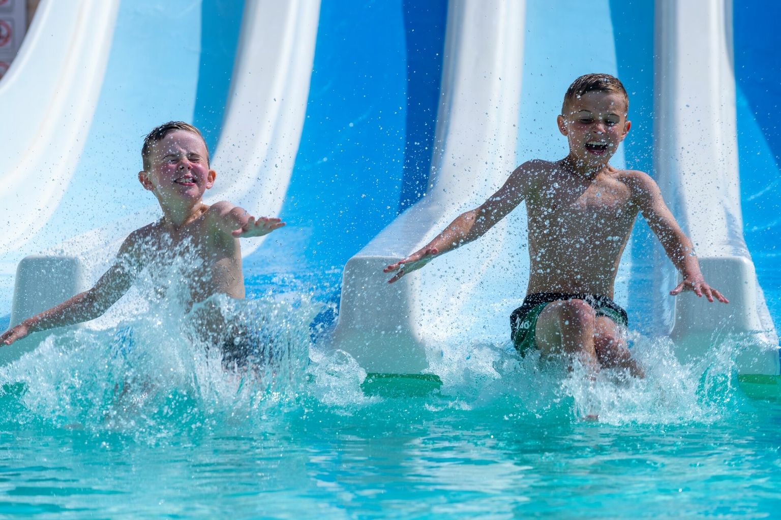 At Le Carbonnier, two kids dive into a swimming pool from water slides, giggling in the bright, open environment.