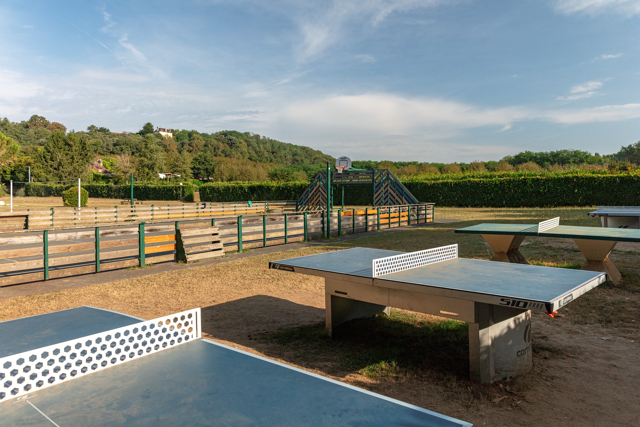 At Marvilla Parks Domaine de Soleil Plage, ping pong tables remain idle in an outdoor sports area, encircled by hedges and distant verdant hills.
