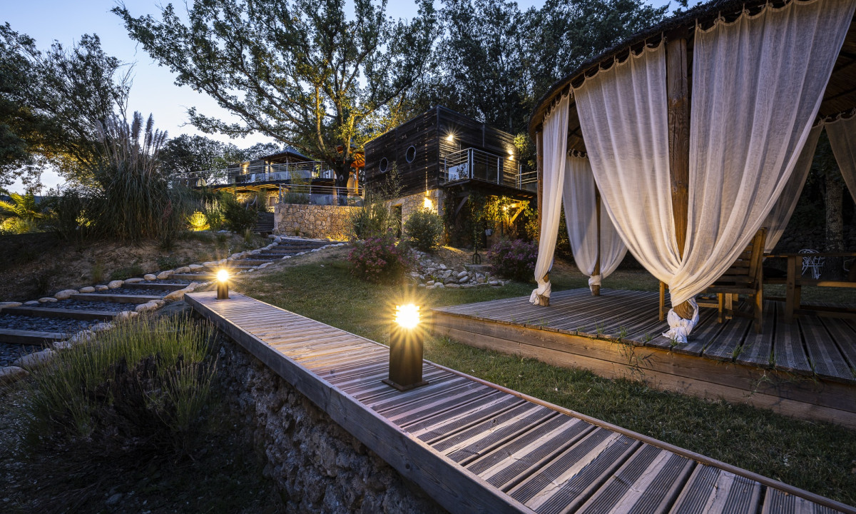 A wooden pavilion with billowing curtains stands illuminated by pathway lights near modern cabins and trees at Domaine de Sévenier