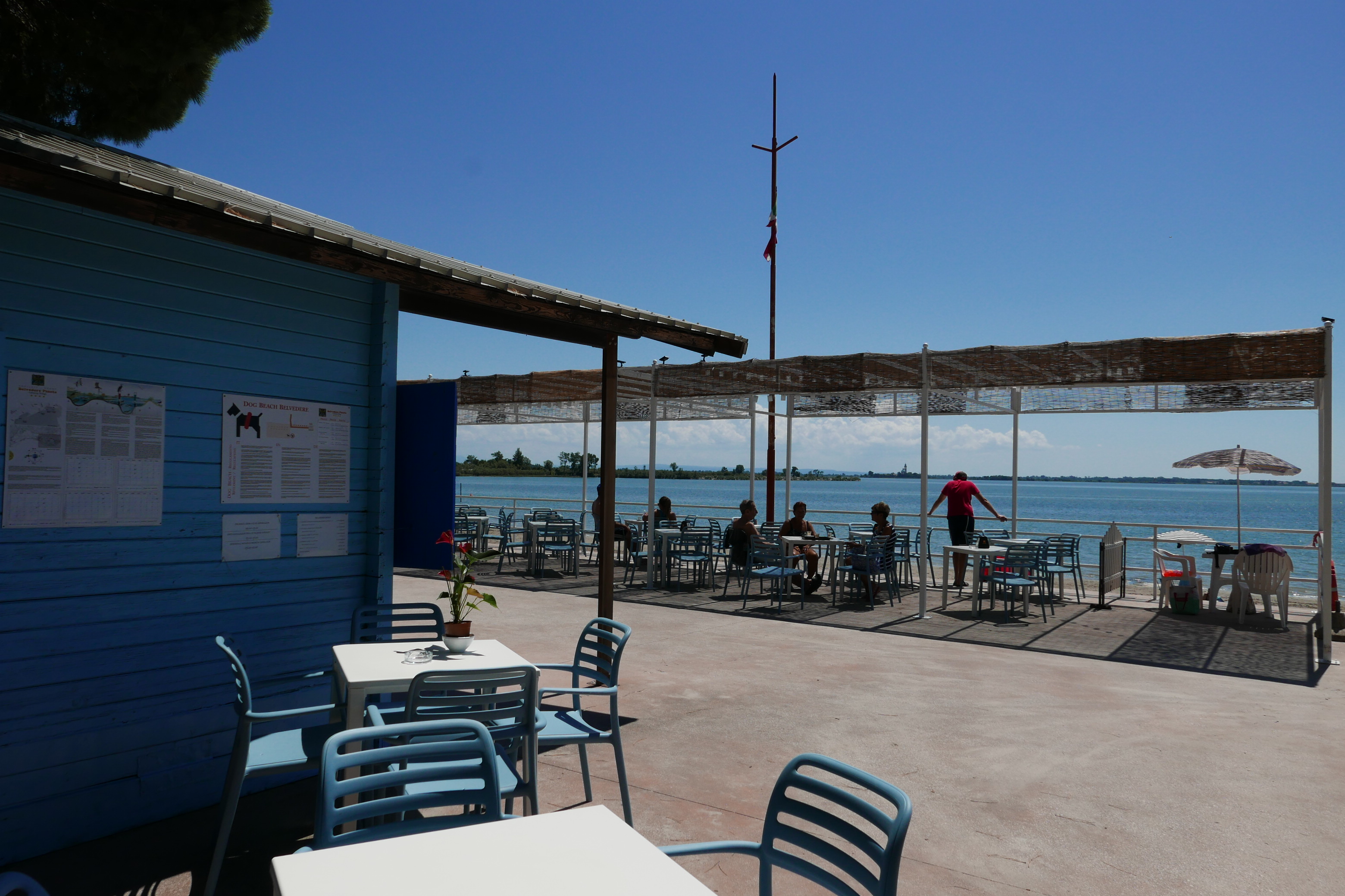 Chairs and tables arranged near a blue building, people sit and stand, seaside backdrop on sunny day at Belvedere Pineta