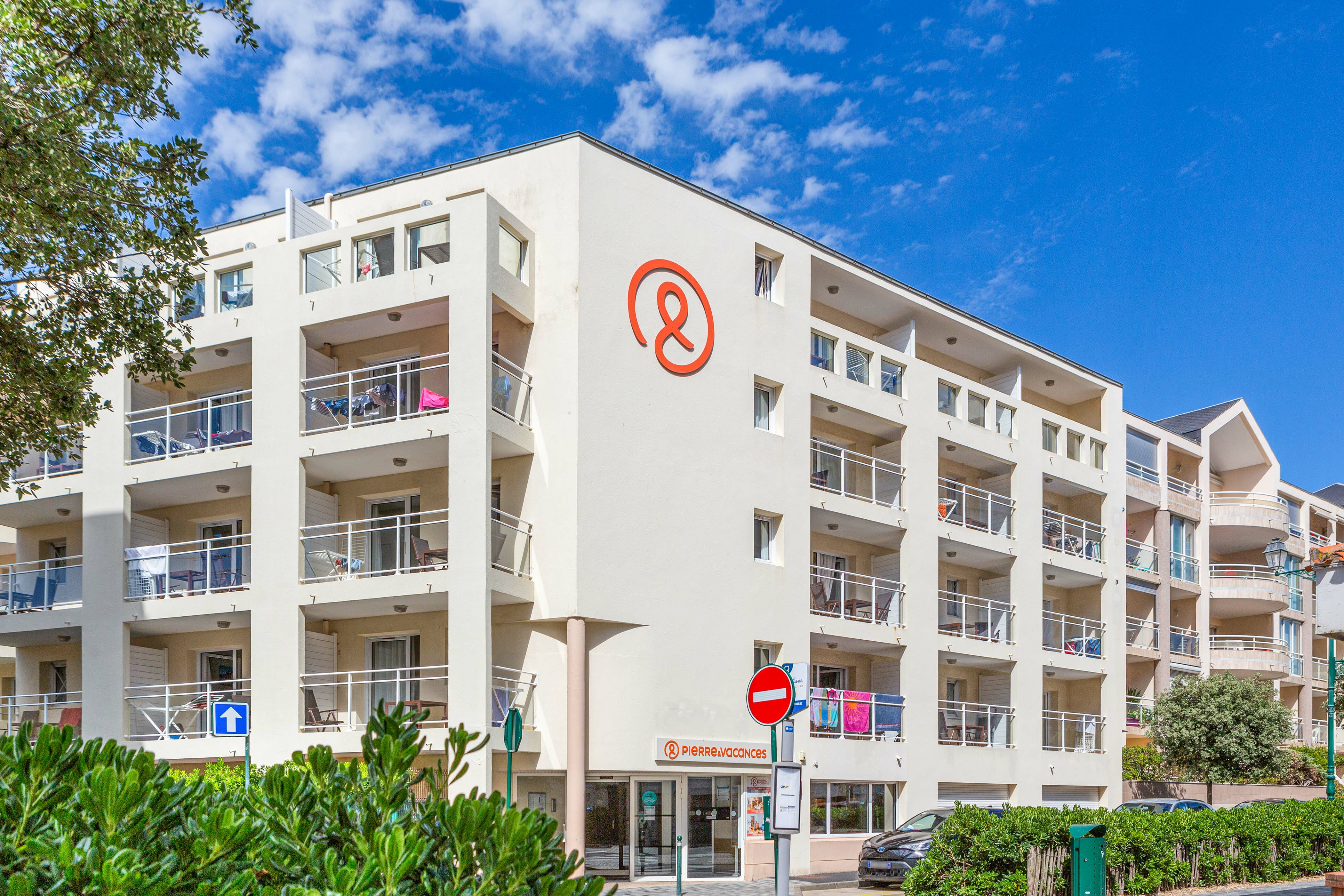 Multi-story apartment building with balconies, under a clear blue sky, surrounded by green foliage and a few parked cars at La Baie des Sables
