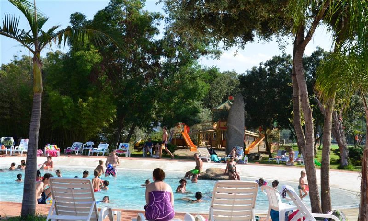 At Ultima Spiaggia, individuals are swimming and relaxing near a spacious outdoor pool, with trees, slides, and lounge chairs visible in the background.