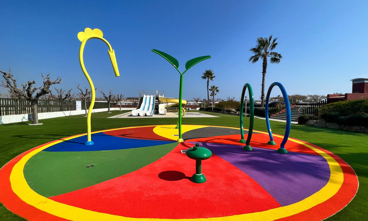 Colorful playground structures stand on vibrant rubber flooring, surrounded by grassy area and clear skies at Resort Els Pins