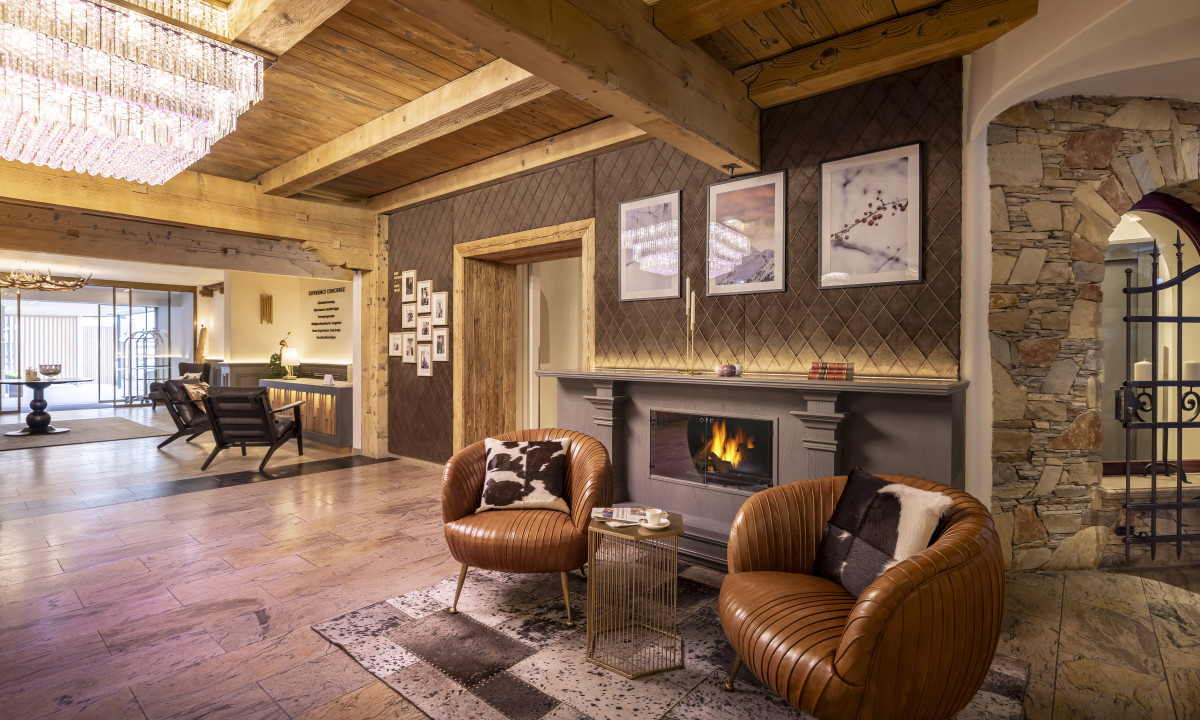 Chairs sitting by a lit fireplace in a cozy, warmly decorated rustic lounge with wooden beams at Kesselspitze Valamar Collection Hotel