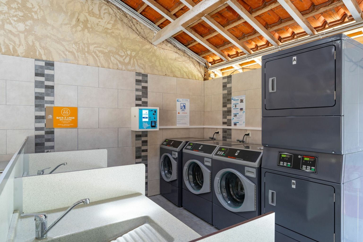 At Le Carbonnier, washing machines and dryers function in a tiled laundry area featuring a rustic ceiling.