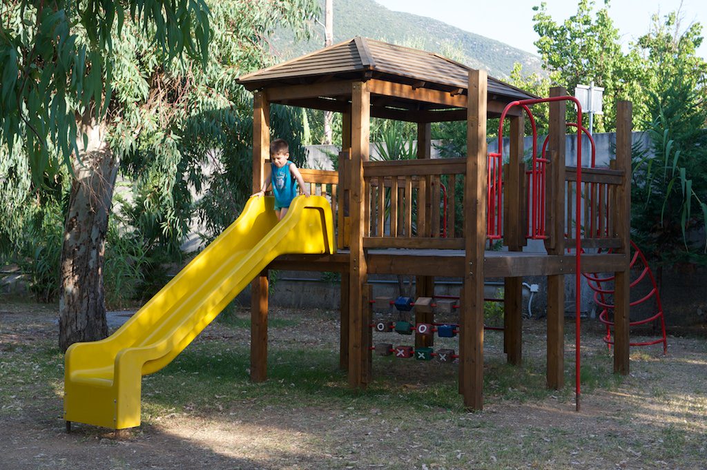 At Karavomilos Beach, a child glides down a golden slide connected to a timber play structure, encircled by trees.