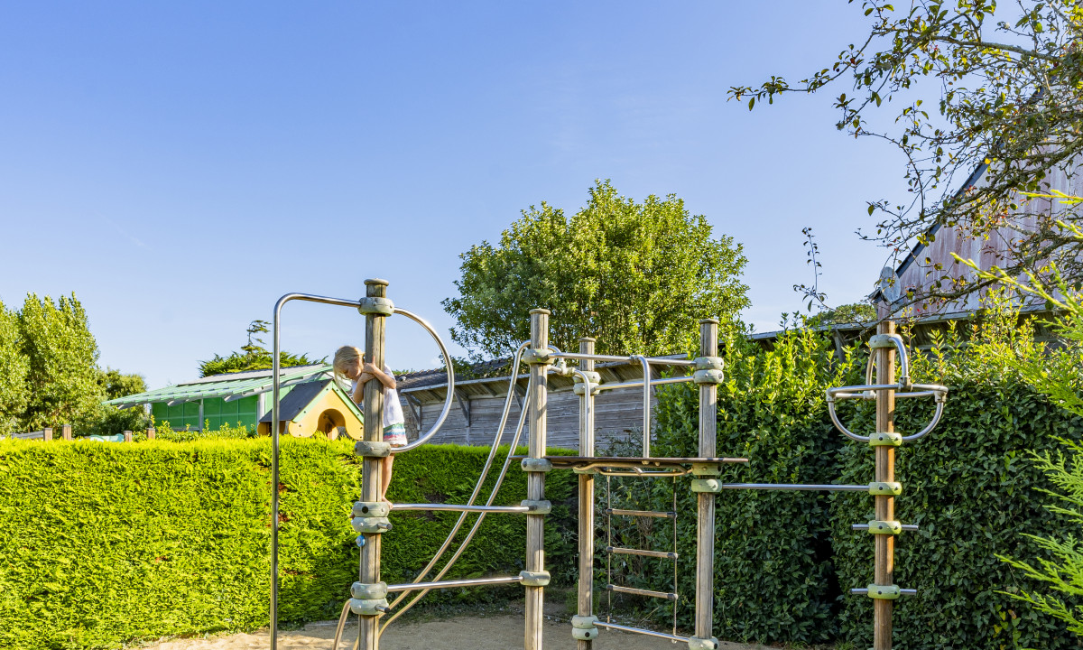 At De la Plage, a child is scaling a play structure, encircled by lush hedges and trees, beneath a clear azure sky.