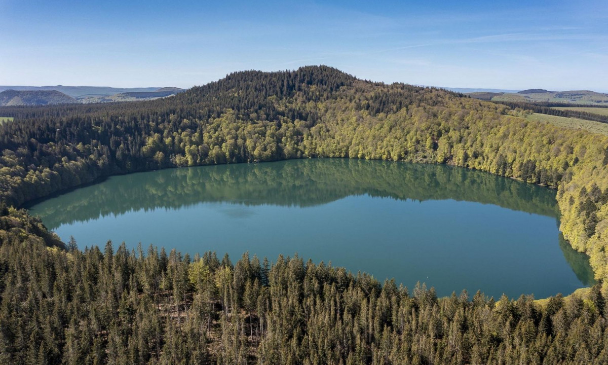 A serene, circular lake reflecting lush, surrounding forest with a high, forested hill in the background at La Ribeyre