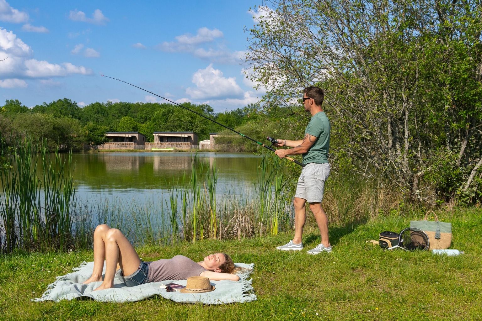 At Le Carbonnier, an individual is angling by the lakeshore while another person relaxes on a blanket, encircled by trees.