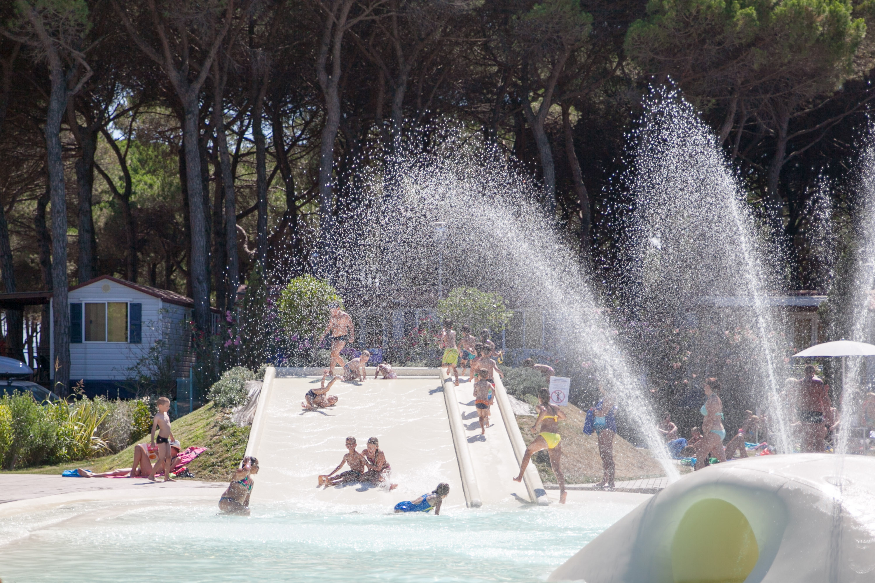 Children and adults enjoy water slides surrounded by misty water sprays and trees at Pineta Sul Mare Camping Village
