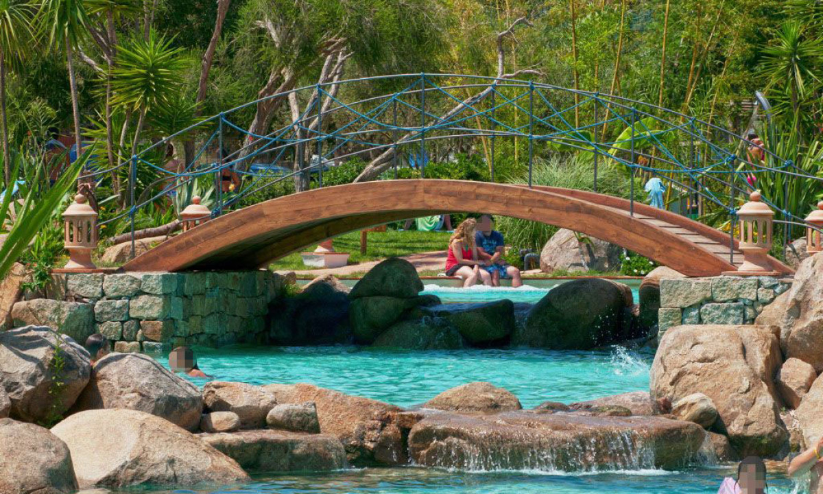 At Ultima Spiaggia, a timber bridge spans a stony stream, with individuals and vibrant foliage in the backdrop.