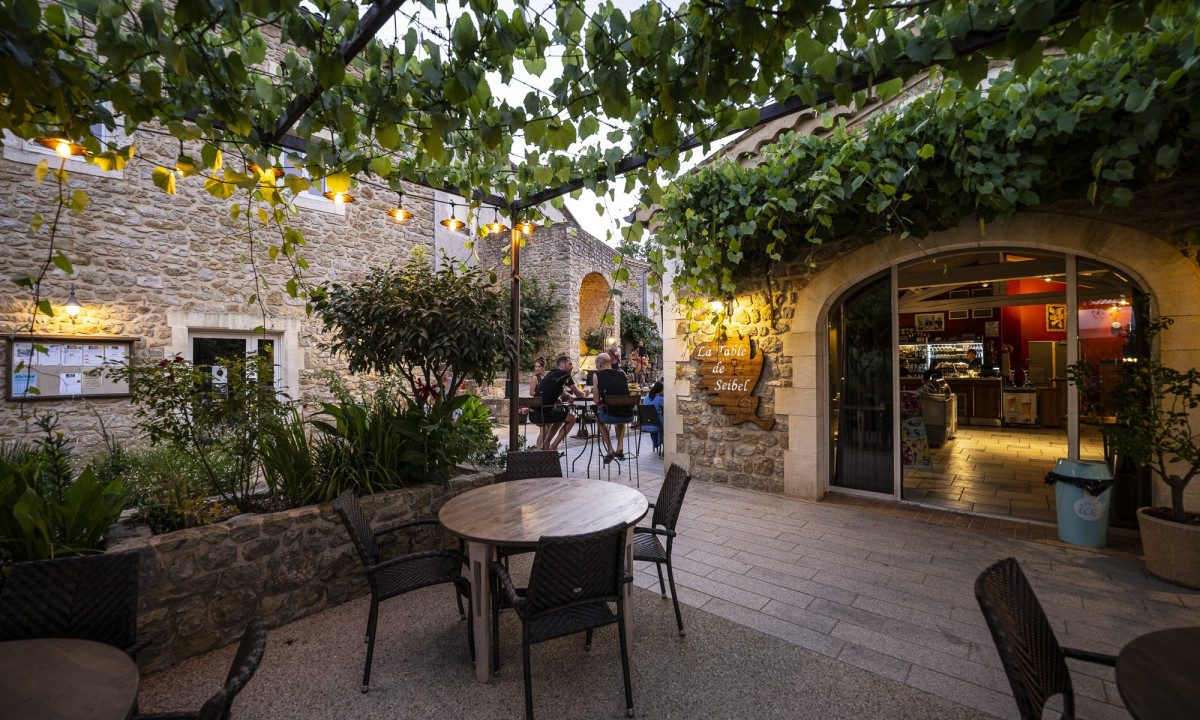 Outdoor dining tables under leafy pergola with string lights, adjacent to rustic stone buildings at Domaine de Sévenier