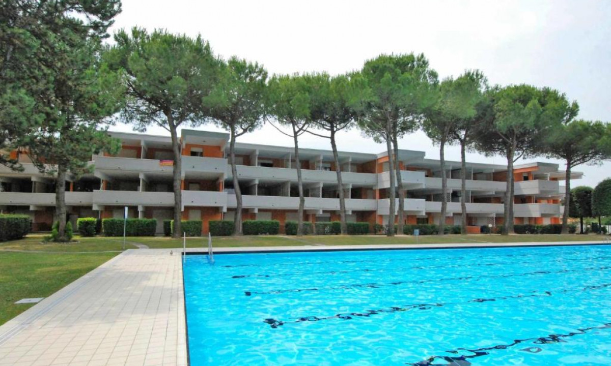 Outdoor swimming pool reflecting clear water, surrounded by a tiled deck, adjacent to a multi-story building with balconies and tall trees at Solarium