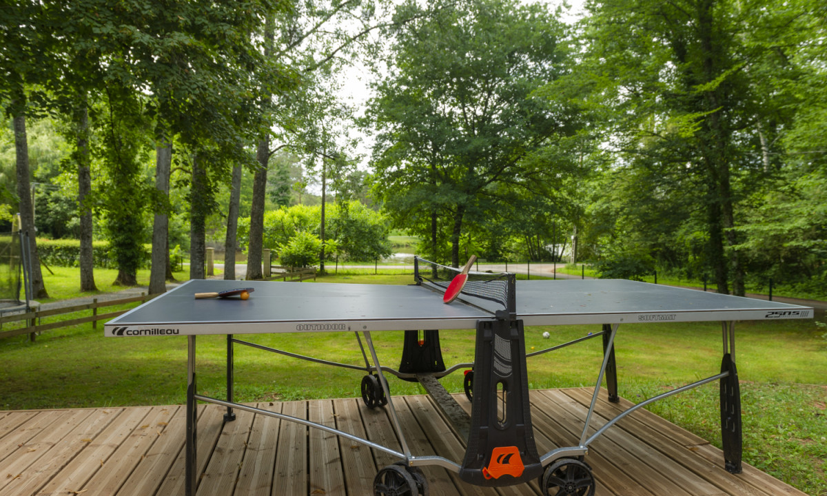 At Coucou, a ping pong table with paddles is situated on a wooden deck, encircled by vibrant green trees and a grassy area.