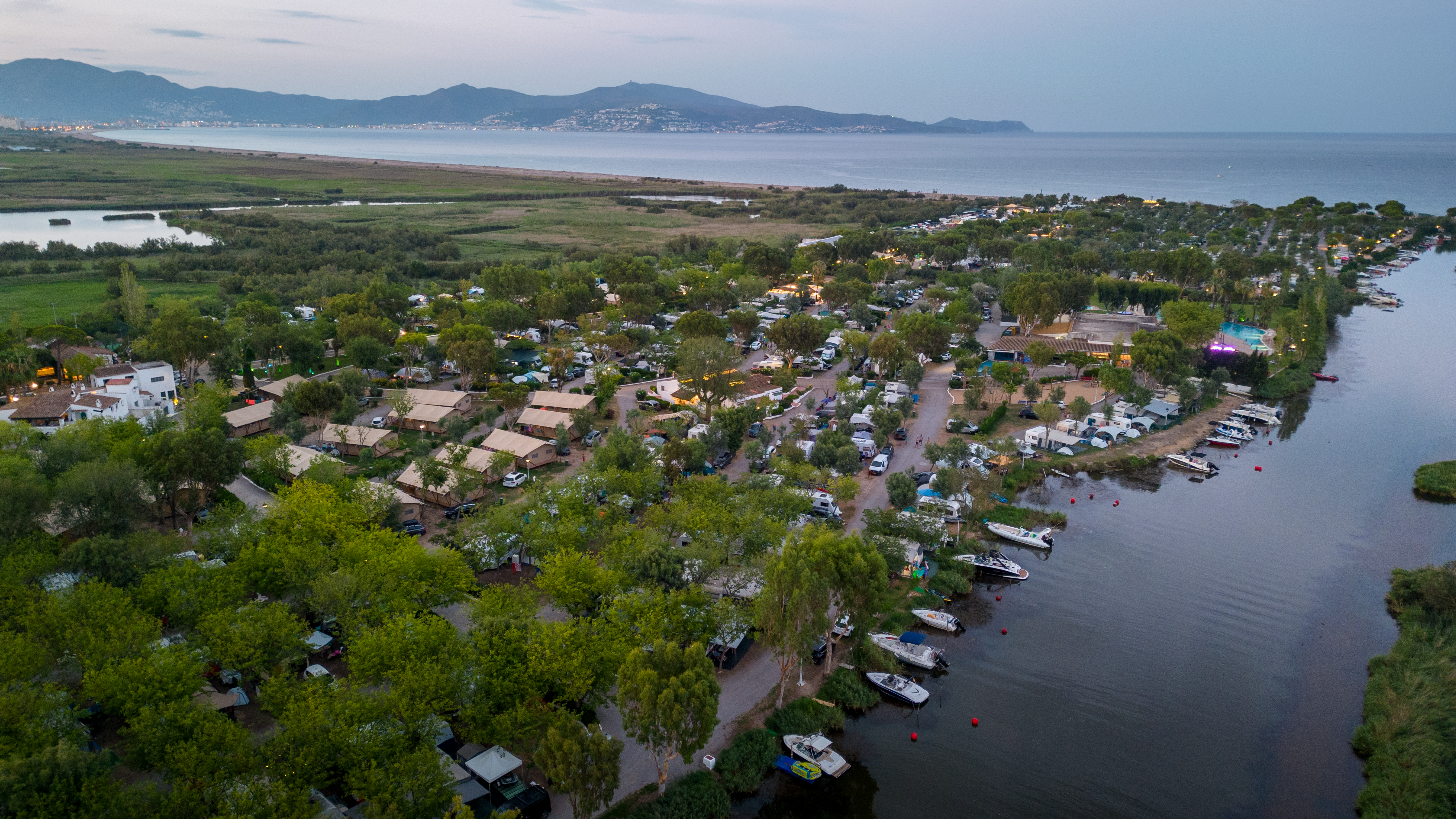 At Nautic Almata, tents and caravans are dispersed among trees, adjacent to a river with moored boats, providing a view of distant mountains and coastline.