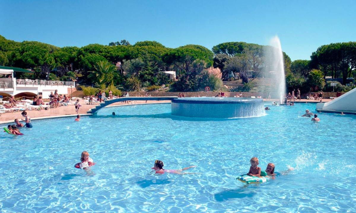 People swim and lounge around a large outdoor swimming pool with a central fountain, surrounded by greenery at Calella de Palafrugell