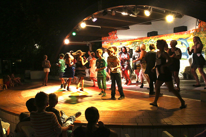 Group of people dancing on an outdoor stage, under colorful lights with seated audience at Belvedere Pineta