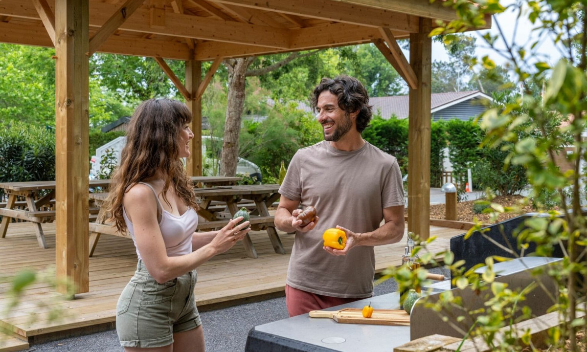 Two people preparing vegetables together under a wooden pavilion, surrounded by picnic tables and greenery at Maguide