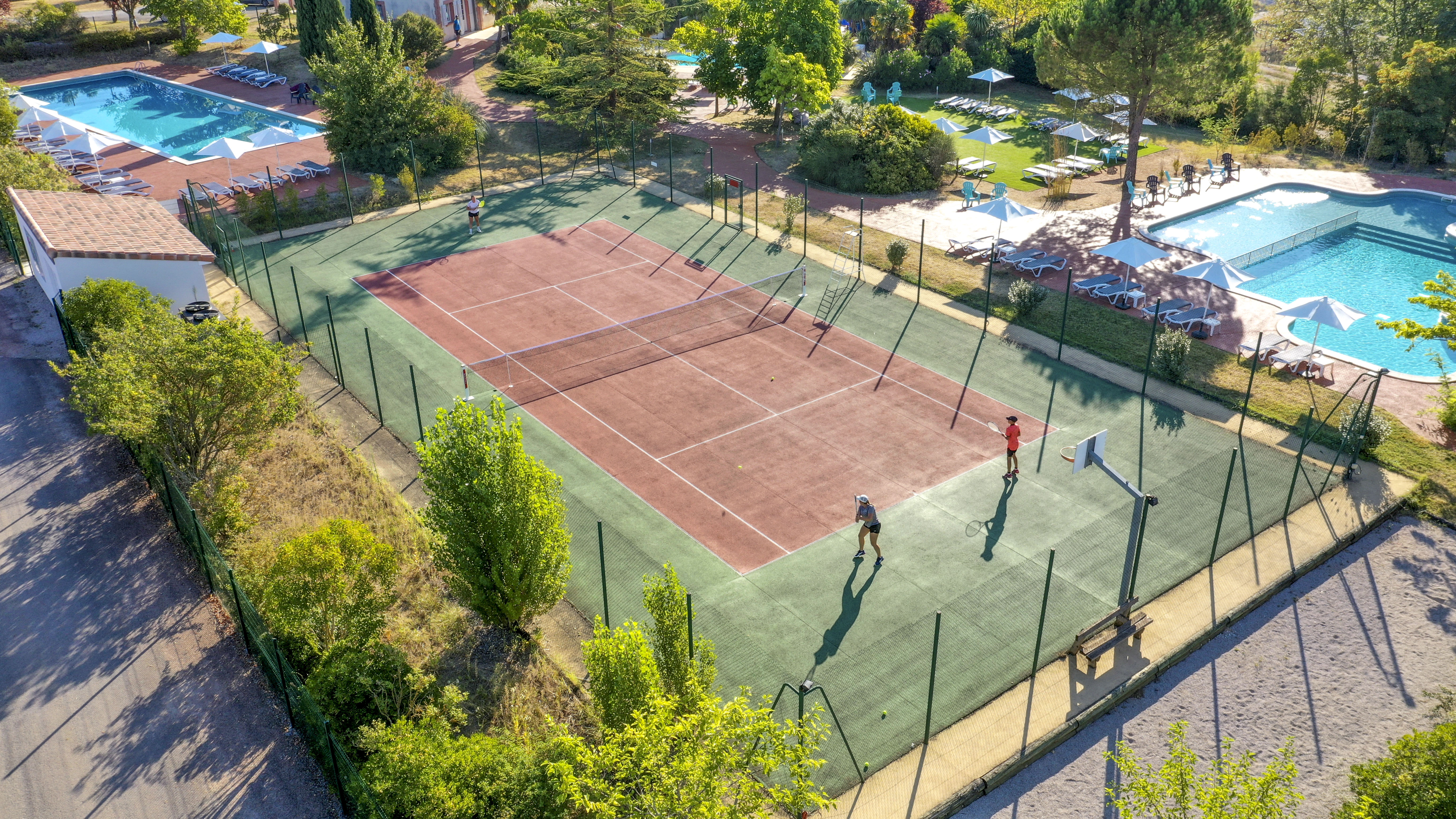 At Domaine du Koukano, individuals engage in tennis on a court next to swimming pools and sunbathing zones, encircled by trees.