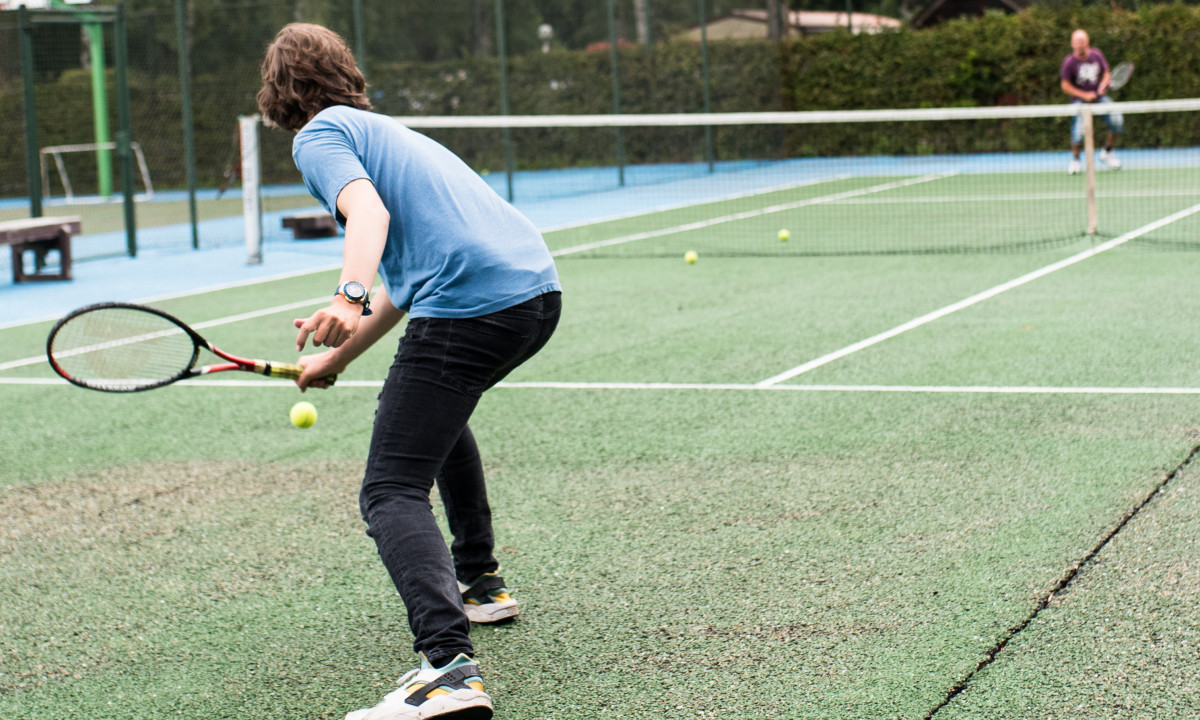 At het Veen, an individual is playing tennis, striking a ball with a racket on a green court, while another person is in the background.