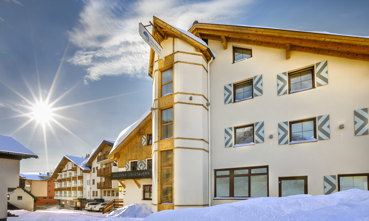 A snow-covered hotel with a wooden facade and bright sun overhead, set in a mountainous winter landscape at Valamar Obertauern Hotel