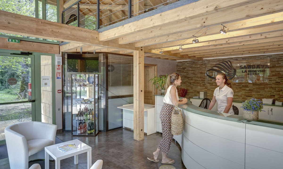 Two women converse at a reception desk in a modern, wooden-themed lobby with chairs and a glass bottle display at La Ribeyre