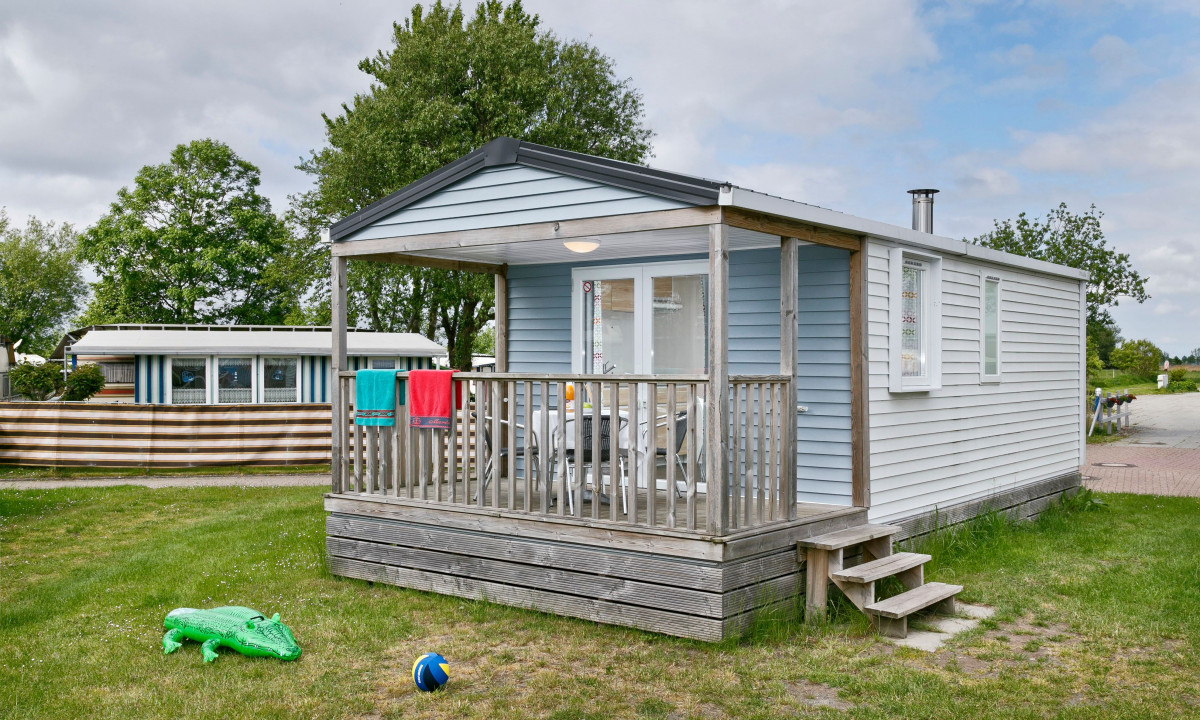 At KNAUS Campingpark Eckwarderhörne, a quaint blue cabin is situated on a grassy patch, with towels draped over the porch railing and children's playthings strewn about.