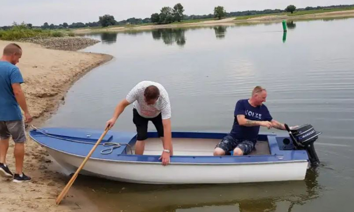 At Elbeling, three individuals ready a small motorboat on a tranquil lakeside, featuring a sandy shore and distant trees.