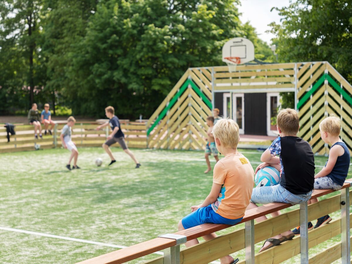At De Twee Bruggen, kids are playing soccer while others observe on a small artificial pitch, encircled by greenery.