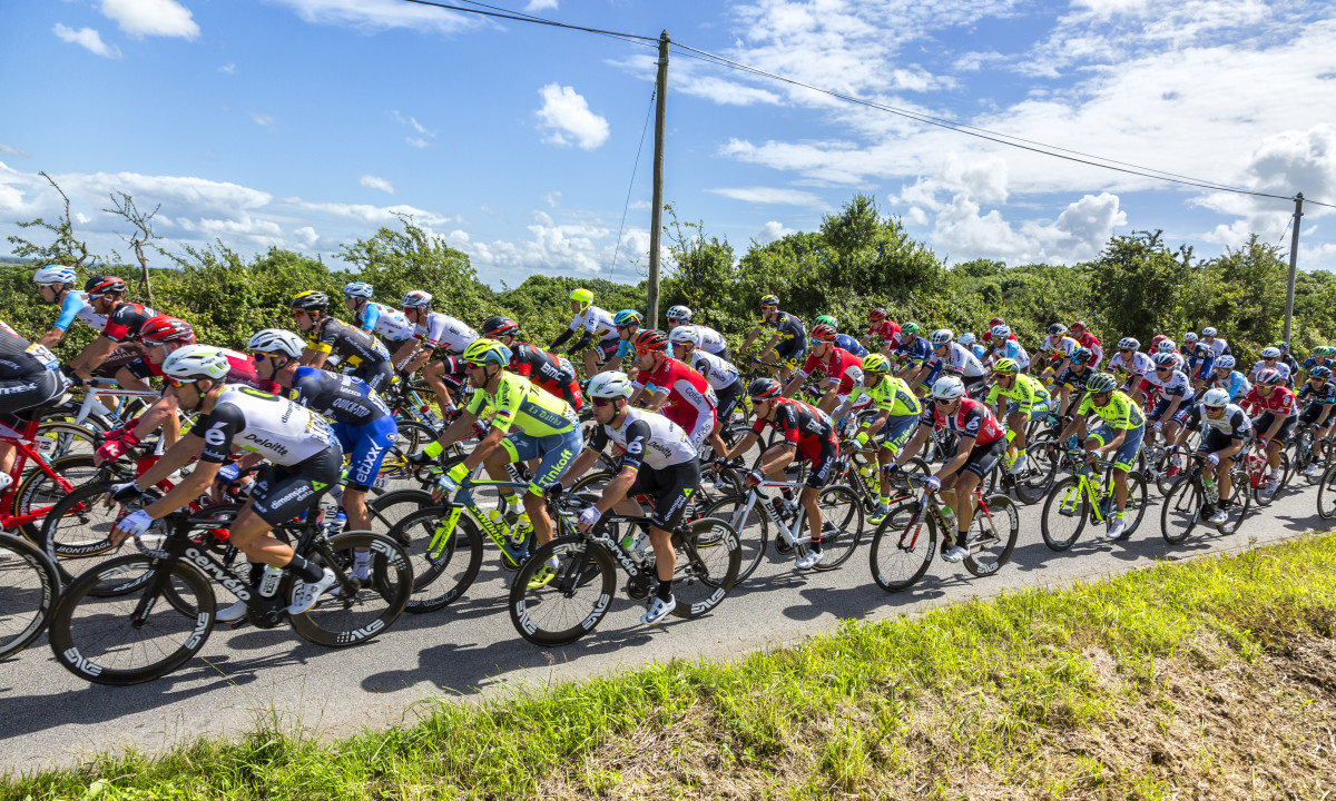 At Urban Gardens Gent, cyclists compete in a closely knit pack on a countryside road beneath a clear azure sky.