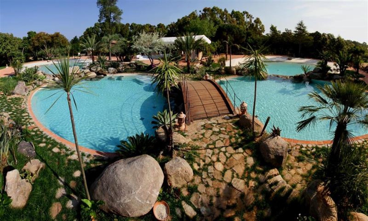 At Ultima Spiaggia, a bridge extends over a lagoon-style swimming pool featuring a stone walkway, encircled by vibrant foliage and trees.