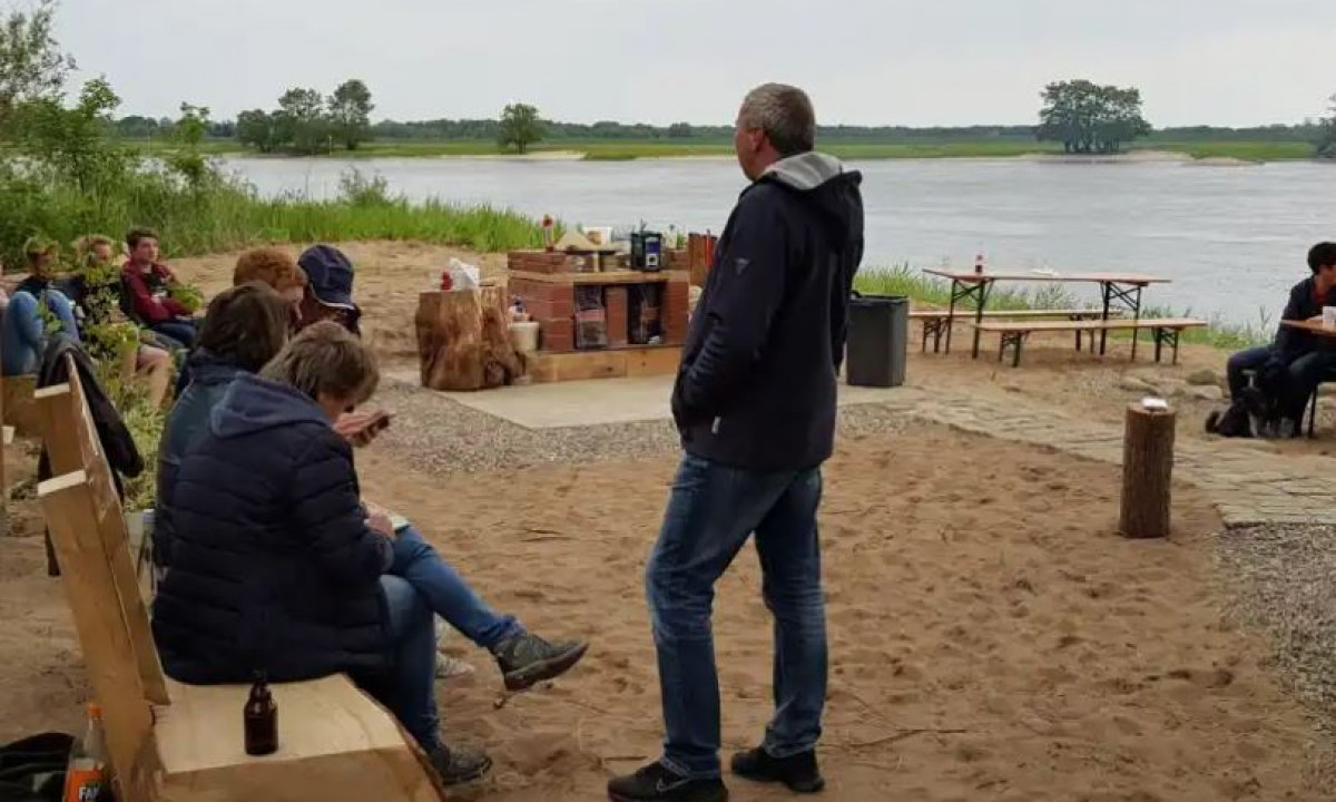 At Elbeling, individuals congregated around a fire pit, with some perched on wooden benches, close to a body of water.