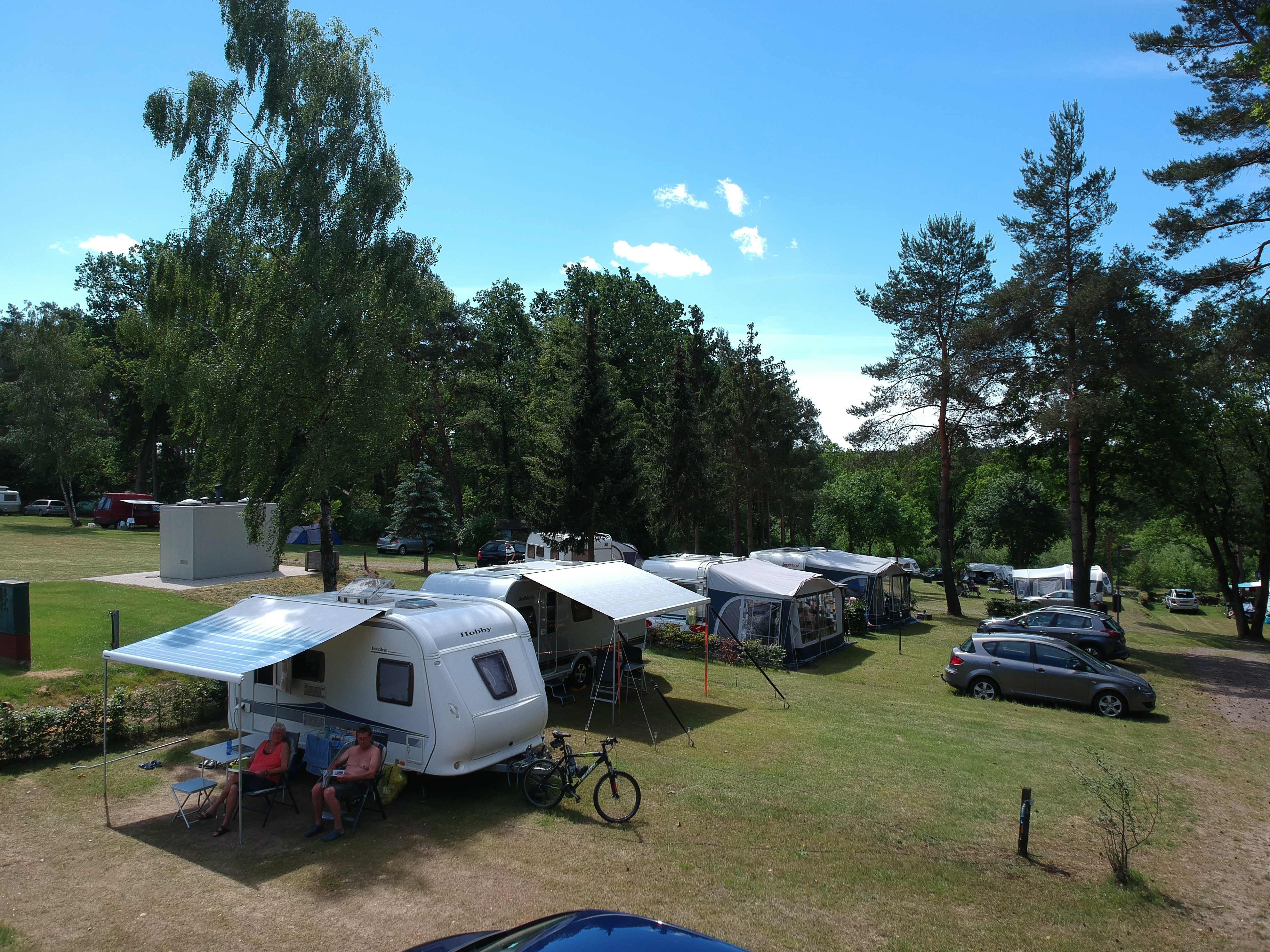 At Sonnenberg, a caravan with an awning is parked, with two individuals seated outside, encircled by trees and parked vehicles.
