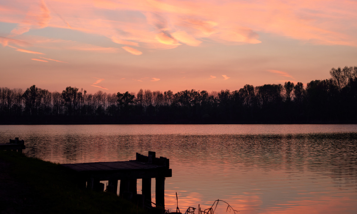 At Urban Gardens Gent, a wooden pier mirrors in tranquil lake waters beneath a vivid sunset sky, bordered by shadowy trees.